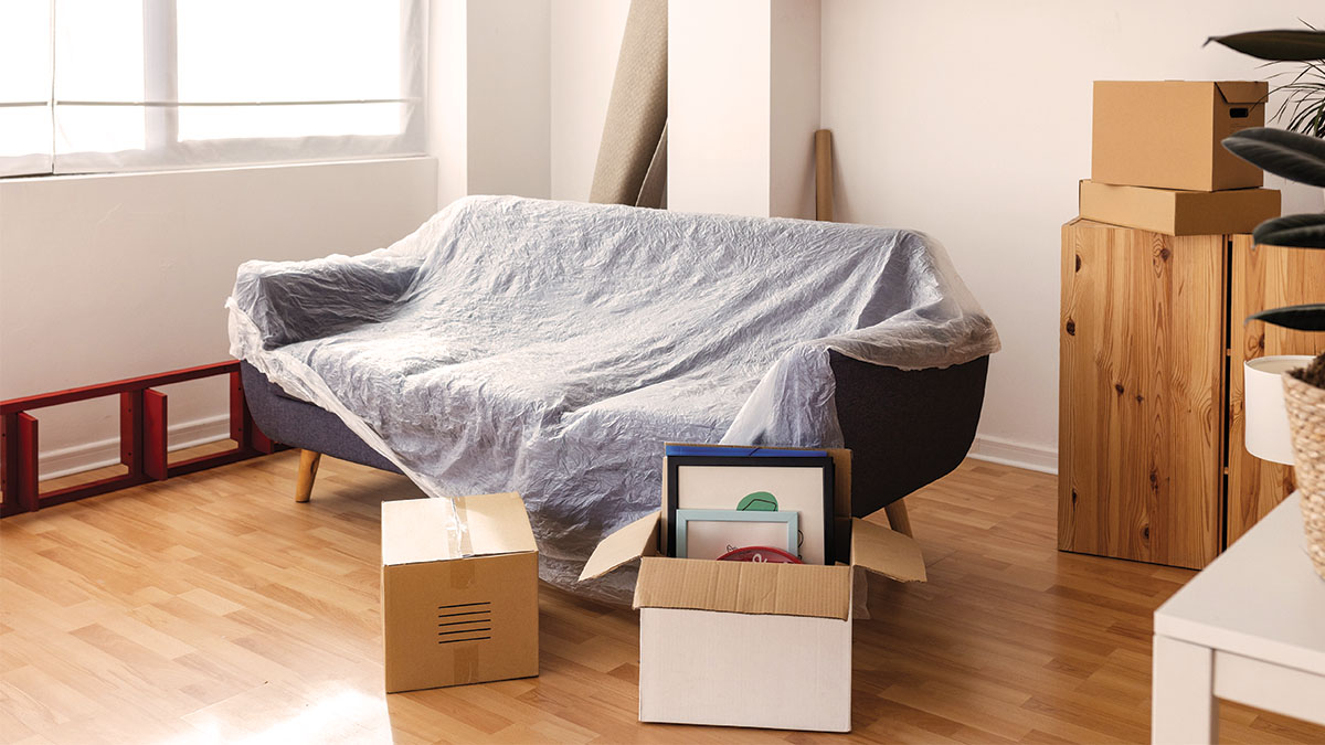 A couch with a mocing cover on it and moving boxes around it A couch with a mocing cover on it and moving boxes around it