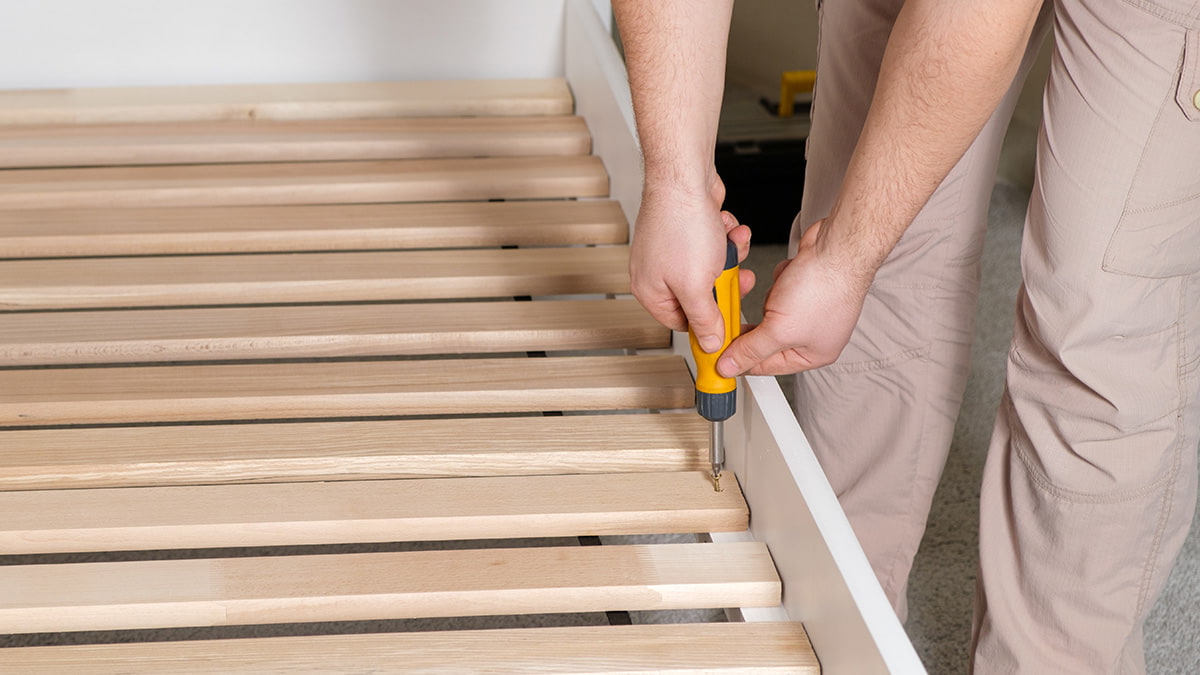 How to take apart a bedframe Person using yellow screwdriver to remove wooden bedframe slats